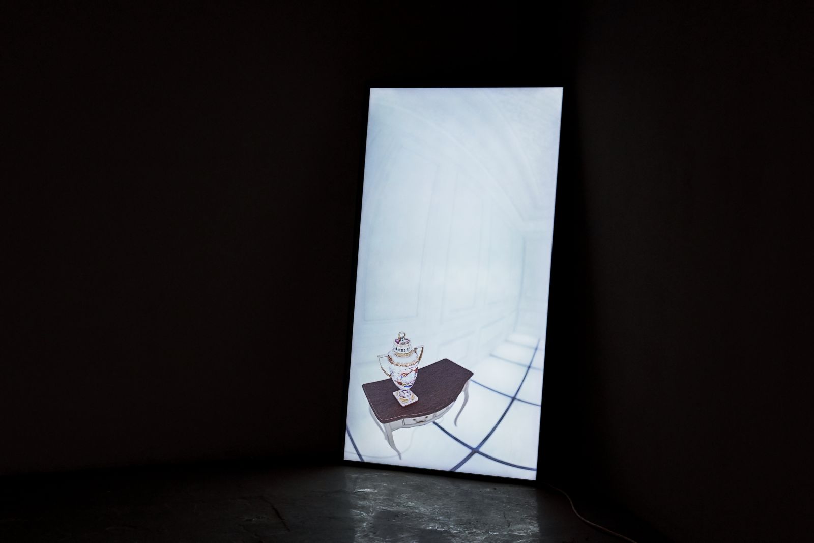 A vertically positioned screen in leaning against the wall in a pitch black space, on the screen is a white room with a baroque side table and a vase